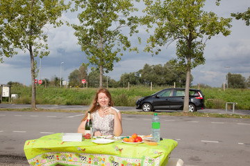 Roadside picnic. A lonely middle-aged woman drinks wine at a table by the side of the road.Concept...