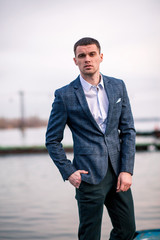 Young man businessman in jacket stands on pier against boat background