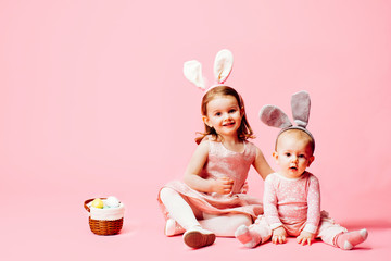 Portrait of a little toddler girl and a baby with bunny ears and easter eggs basket, on pink