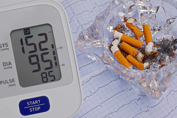 automatic blood pressure meter and ashtray with cigarette butts on cardiogram graph background