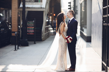 Naklejka premium Elegant wedding couple walking in a summer city. Man in a blue suit. Woman in a white elegant dress with bouquet of roses
