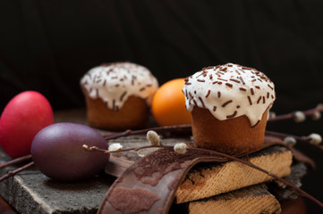 A  two small Easter cake and dyed  eggs, and a willow twig on a dark wooden background. Close up. Selective focus.