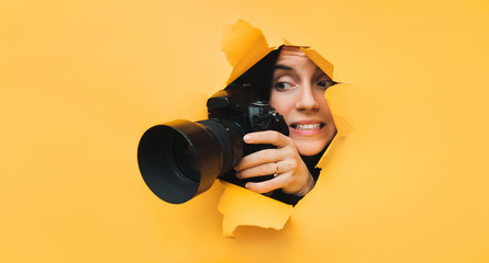 A young paparazzi girl holds a reflex camera and looks through a torn hole in yellow paper. The...