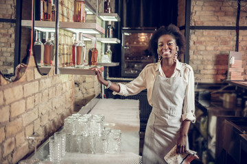 Smiling bewitching dark-haired African-American waitress standing near bar