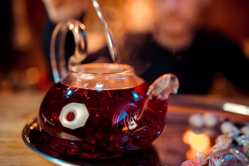Red tea in a teapot on a stand with a candle on wooden bar counter.