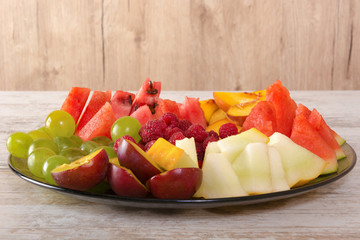 Fruit mix. Peach, grapes, apple, watermelon, melon, raspberry, plum. Pieces of fruit on a plate lie on a white wooden background. Copy space, space for text.