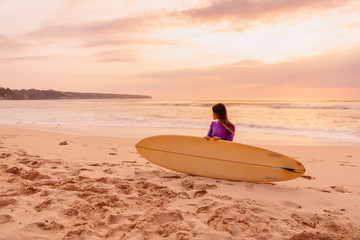 Surfer girl with surfboard on a beach at sunset or sunrise.