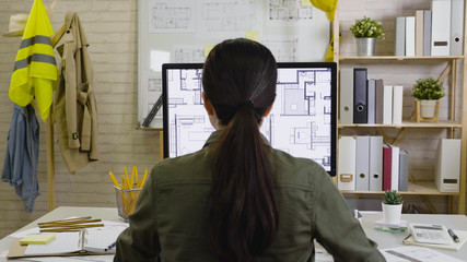back view of young asian woman employee looking on monitor with blueprint architect construction project working in morning in office. korean girl interior designer correcting floor plan deadline.