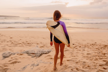 Surfer girl with surfboard on a beach at sunset or sunrise.