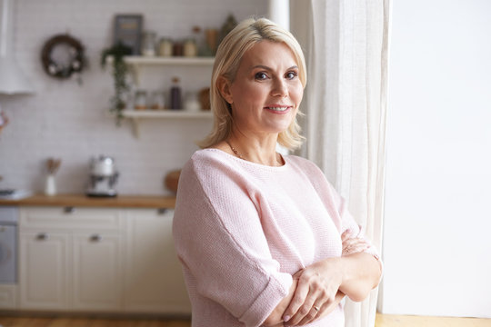 Horizontal indoor view of beautiful friendly looking European woman in her forties spending day at home, standing by window with arms crossed on her chest, having happy cheerful facial expression