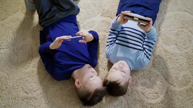Boys lying on the floor on a light carpet and playing games on mobile phones at home. Top view of two kids playing together and helping each other in games.