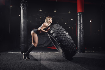 Sports woman is engaged in workout with heavy tire in dark gym