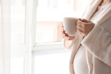 Fototapeta na wymiar Pregnant woman with cup of hot tea standing near window at home
