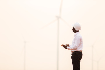 African engineer wearing white hard hat standing with digital tablet against wind turbine on sunny day