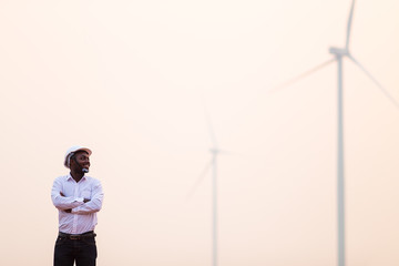 African engineer wearing white hard hat standing with digital tablet against wind turbine on sunny day