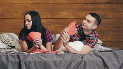 Man holding three paper hearts gives them to his girlfriend and they are kissing passionately on bed. Happy lovely couple lying in bed flirting to one another at home. Close-up