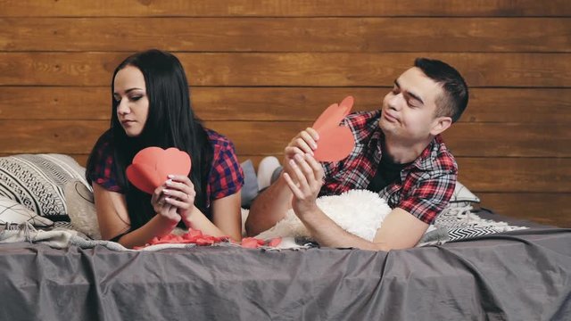 Man holding three paper hearts gives them to his girlfriend and they are kissing passionately on bed. Happy lovely couple lying in bed flirting to one another at home. Close-up