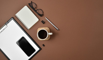 Flat lay, top view office table desk. Workspace with blank clip board, notebook, office supplies, spectacles, pen, mobile phone and coffee cup on brown background
