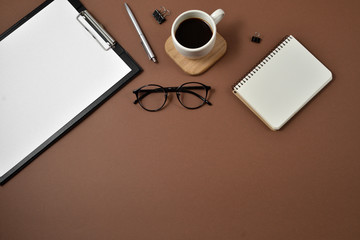 Flat lay, top view office table desk. Workspace with blank clip board, notebook, eyeglasses, office supplies, pen and coffee cup on brown background. Blog concept