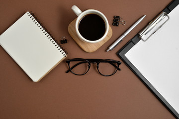 Flat lay, top view office table desk. Workspace with blank clip board, notebook, eyeglasses, office supplies, pen and coffee cup on brown background. Blog concept