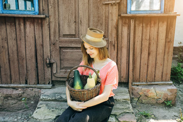 Beautiful girl in old rustic clothes and straw hat is sitting on stairs near the house. Summer day on farm barn yard. Natural woman with basket of zucchini courgette from home garden. Stylish vintage.