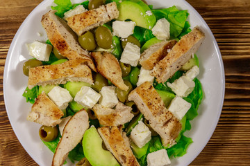 Tasty salad with fried chicken breast, green olives, feta cheese, avocado, lettuce and olive oil on wooden table. Top view