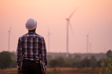 African engineer wearing white hard hat standing with digital tablet against wind turbine on sunny...