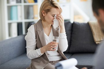 Sad and depressed young woman wiping her tears and having glass of water while sitting on couch in psychologist office