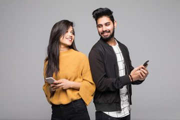 Beautiful young indian couple holding mobile phones and standing back to back against grey background