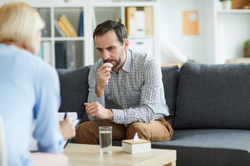Pensive middle aged patient sitting on couch and bending over table during consultation with his...