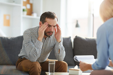 Tense and exhausted man trying to explain his troubles to psychologist while sitting on couch in front of her