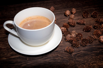 white cup of coffee on wood table and some pine cones on the background.