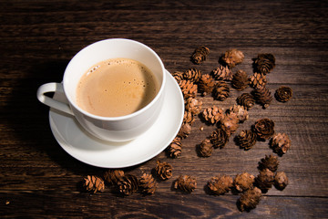 white cup of coffee on wood table and some pine cones on the background.