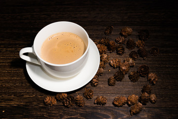 white cup of coffee on wood table and some pine cones on the background.