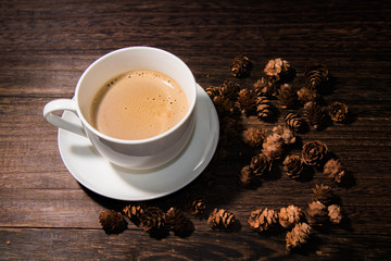 white cup of coffee on wood table and some pine cones on the background.