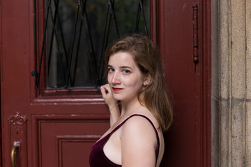 Fototapeta premium Medium horizontal portrait of exquisite blue-eyed young woman in velvet tank top with small smile turning while standing against dark red door