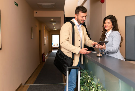 Young Business Man Check-in In Hotel, Smiling Female Receptionist Behind The Hotel Counter Showing Him Available Rooms On Tablet.