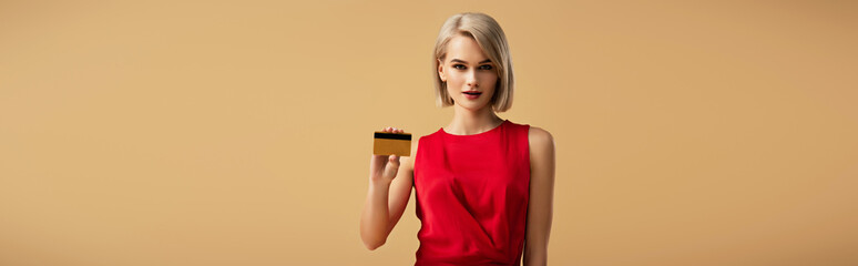 panoramic shot of beautiful young woman in red dress holding credit card isolated on beige