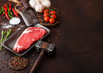 Raw sirloin beef steak in plastic tray with salt and pepper and vintage meat hatchets and hammer on rusty background.Red pepper, tomatoes and garlic. Space for text