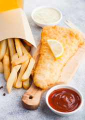 Traditional British Fish and Chips with tartar sauce abd glass of craft lager beer and tomato ketchup on chopping board on white stone table background.