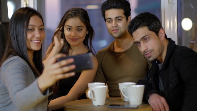 Young friends pose for selfie photos from a mobile phone as they huddle together and pose and try to get a better one over a coffee together on a cafe talking chatting and talking 