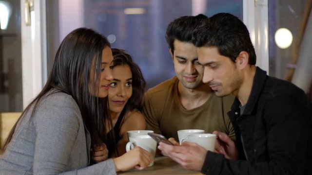Indian young friends intently look at a mobile screen touch screen and look at something as they share, review and criticize the photo over a coffee together on a cafe talking chatting and talking 
