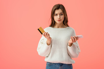 confused brunette girl holding credit card and smartphone isolated on pink