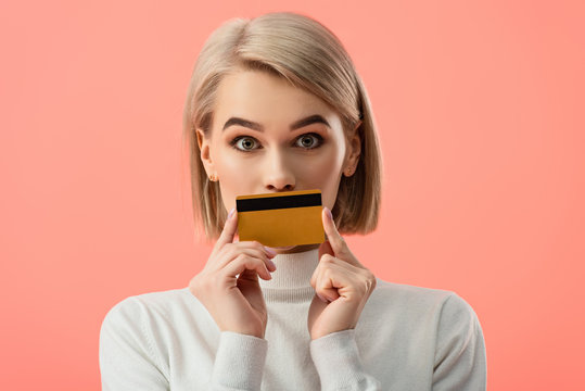 Surprised Blonde Woman Covering Mouth With Credit Card Isolated On Pink