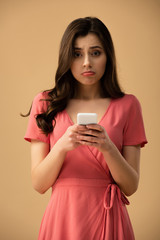 upset brunette girl holding smartphone isolated on brown