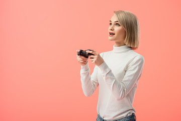 surprised blonde woman holding joystick while playing isolated on pink