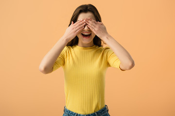 cheerful young woman covering eyes with hands isolated on orange