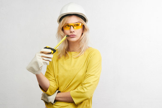 Beautiful blonde engineer in construction helmet, gloves and glasses thinks with pensive face on white background, horizontal shot with copy space