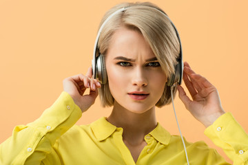 Dissatisfied blonde young woman touching headphones and looking at camera isolated on orange