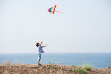 active little caucasian boy in denim shorts controls flying colorful kite in blue sky